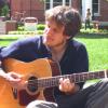 photo of Jon Watts sitting on the grass on a sunny day, playing guitar and singing